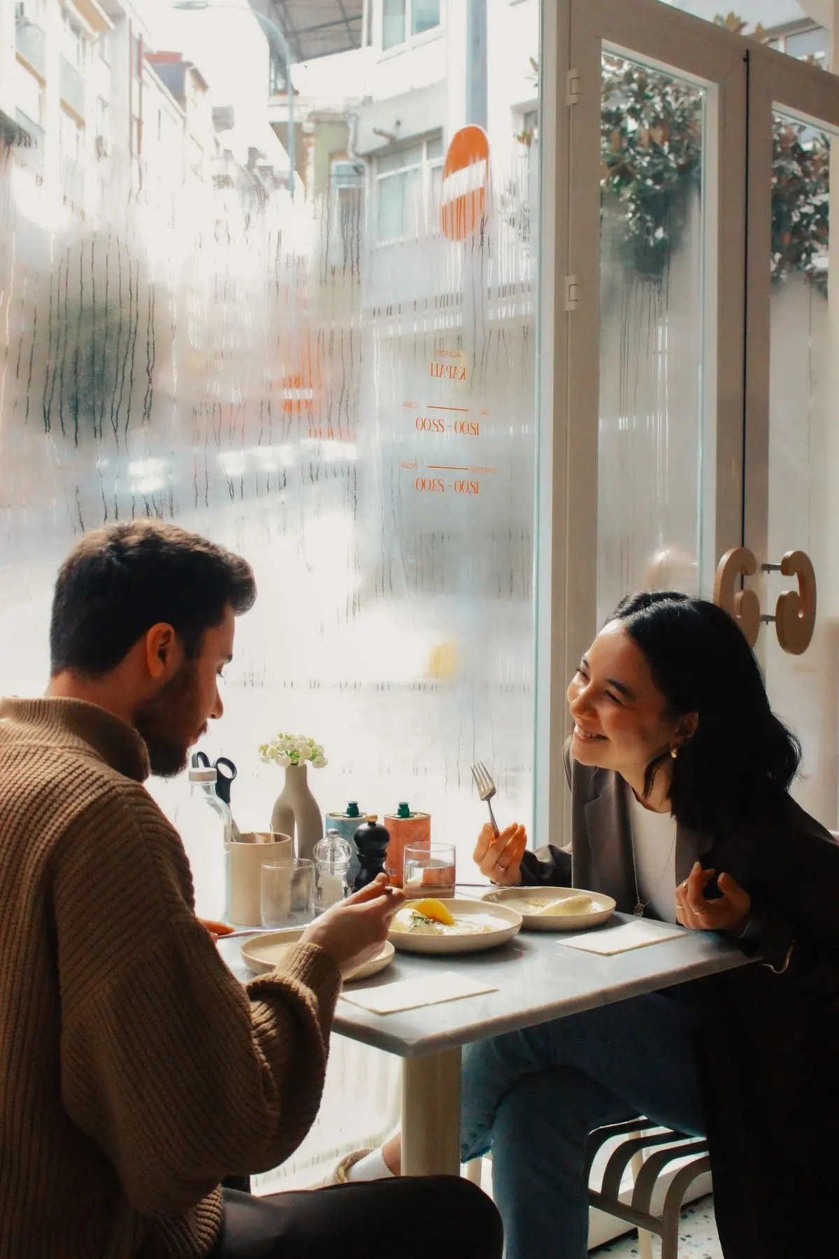 Two people laughing together over coffee by a café window
